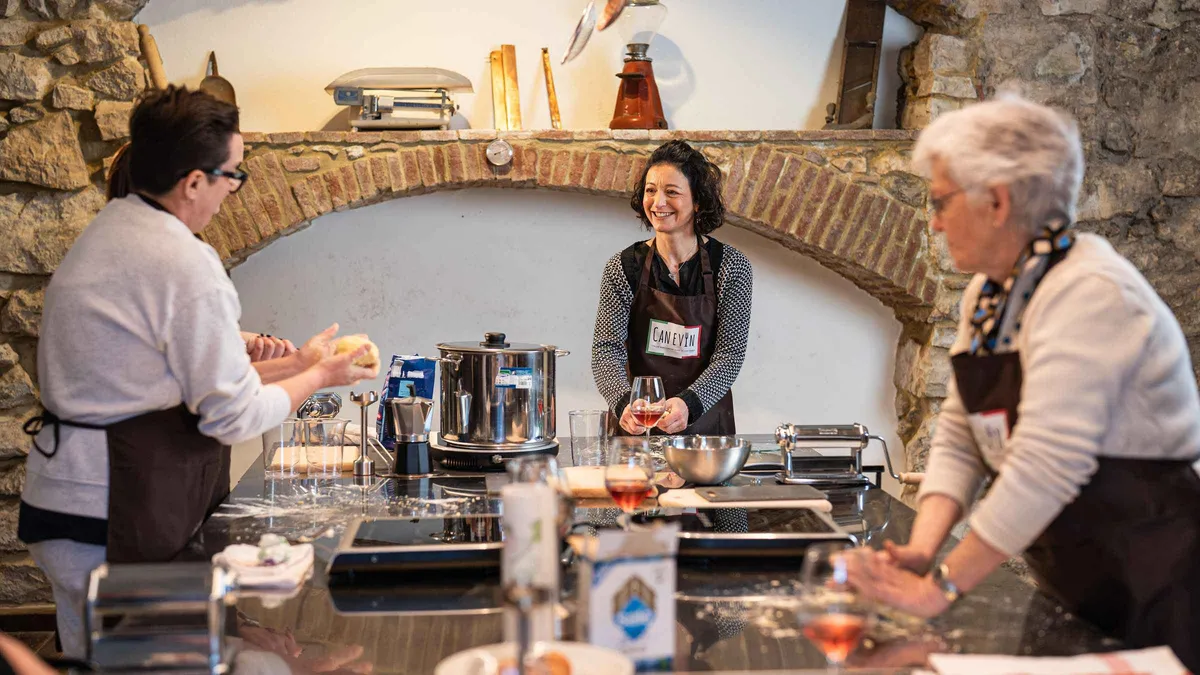 Three women in aprons kneading pasta dough at a kitchen counter under a stone arch