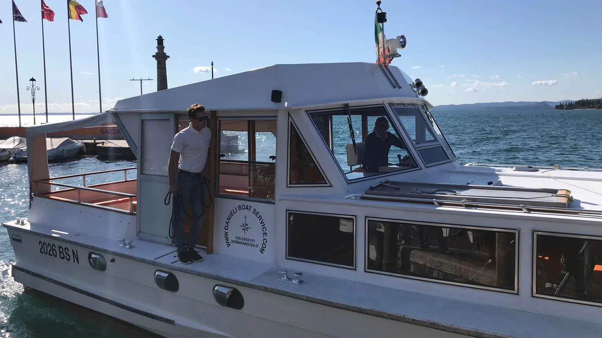 A white motor cruiser with a crew member on deck tied up at a lakeside quay under flags