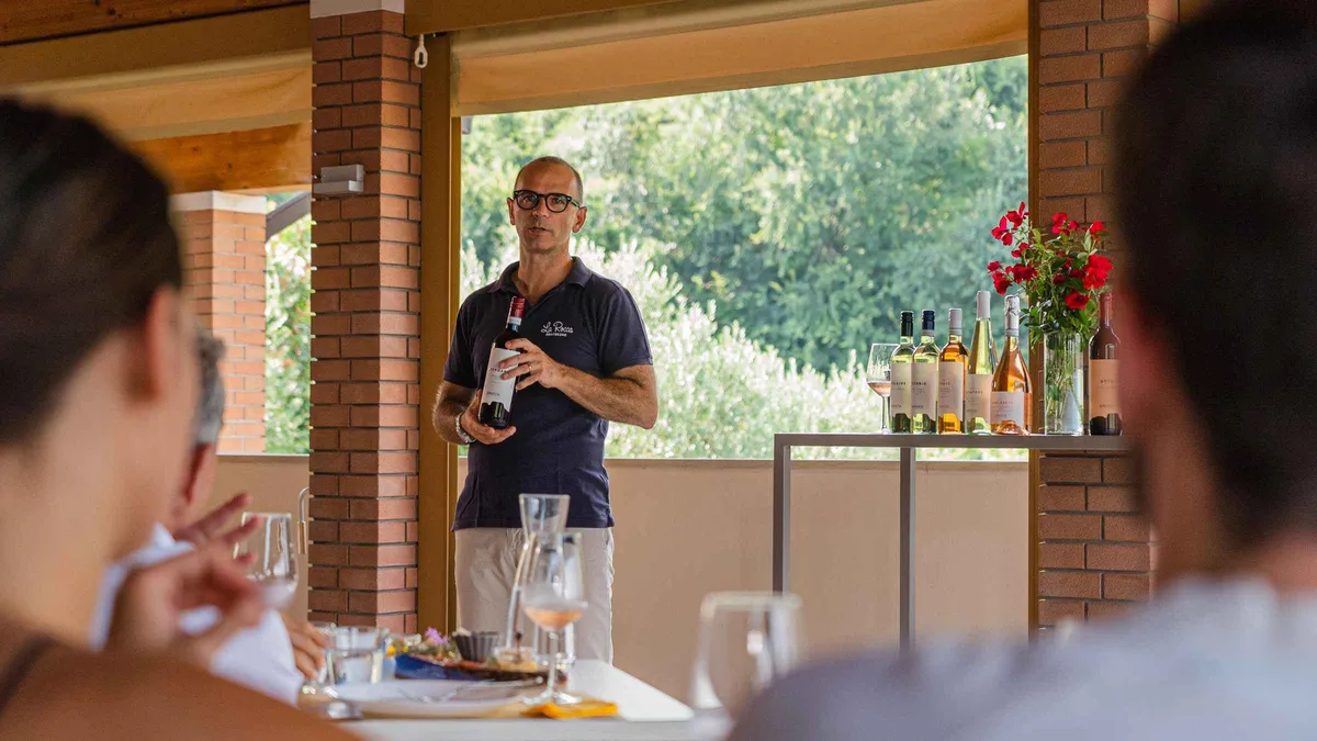 A host pouring wine for guests under a brick porch that opens onto green vines